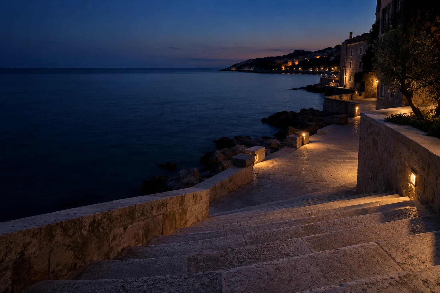 Un chemin au bord de la mer à la tombée de la nuit, avec quelques lumières au loin.